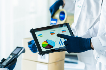 工場や倉庫内でデータ集計用のタブレットとハンディターミナルを持つ白い作業服のスタッフ
